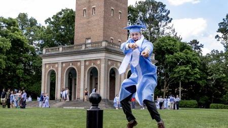 U.N.C graduate in regalia jumping and smiling for photo with Bell Tower seen in background.